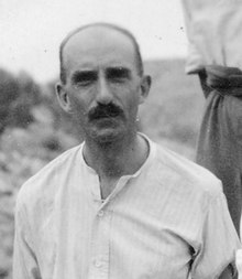 A middle-aged man in an open-necked, Greek-style collarless shirt, with a neat moustache.
