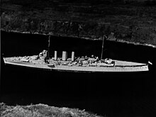 Aerial photograph of a cruiser-size warship sailing slowly through a narrow body of water