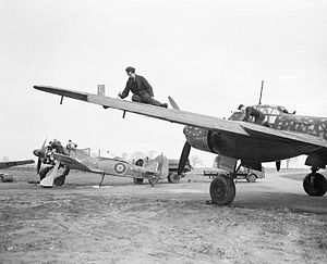 A photograph of a single–engined and a larger, twin-engined aircraft on the ground. Two men are working on the wing of the larger aircraft. The engine–covers have been removed from the smaller aircraft and four men are working on its engine