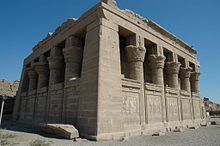 Small stone building surrounded by half-walls and columns with elaborate capitals.