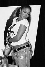 A sideshot of an African-American woman wearing jeans and a T-shirt. She smiles and stares behind herself, down towards the floor.
