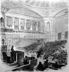 An etching of an ornate two story room filled with people seated in chairs on both the floor and balcony. At upper right is a dark fish shape.