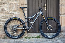 A grey 2021 full suspension mountain bike with a four-bar linkage rear suspension in front of a buildings wall and door.