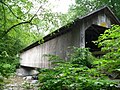 A light brown wooden covered bridge seen from one side of the river it crosses, slightly below the portal. It is surrounded by green tree branches on either side.