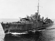 Photograph of a war ship in a body of water. Large cannons can be seen on the deck, along with sailors.