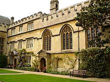 Adjoining buildings with battlements to the side of an area of grass; on the left, regularly-placed windows in a three-storey building, and foliage and flowers growing above and around a stone canopy over the doorway; on the right, a chapel with three arched windows and a stone porch; a bench alongside the chapel, and a tree at the far right