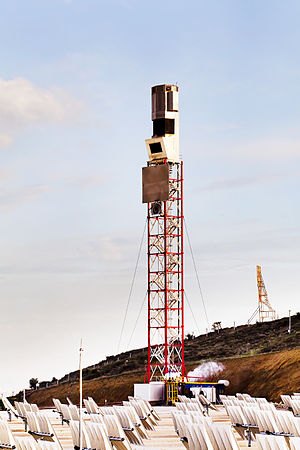 Angled mirrors in the foreground. A guyed metal tower topped with a white box with black rectangle on one side.