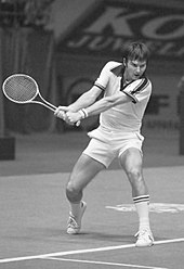 A brown-haired man dressed in a white shirt swings a two-handed backhand
