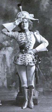 A young white woman standing in costume; she is saluting with one hand, with the other hand at her waist; her costume is corseted and short, with military details, white tights and dark boots; she is wearing gloves and a large hat