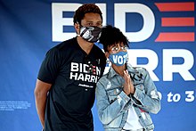 A woman with dark curly hair wearing a face mask with the word "VOTE" on it, clear glasses, a white shirt, and a jeans jacket clamps her hands together as she stands to the left of a man with a short afro wearing a black face mask with a Los Angeles Lakers logo and a dark-colored shirt with the words "BIDEN HARRIS" on it