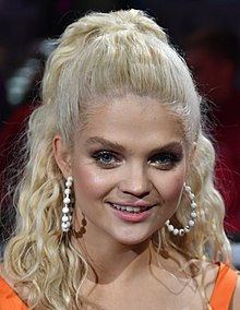 Close-up shot of a smiling Margaret with her hair tied up. She wears white hoop earrings and an orange top.