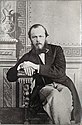 A black-and-white photograph of a full-bearded man sitting with his legs crossed and looking at the viewer while wearing a dark-coloured jacket and light-coloured pants