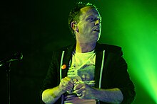 Torquil Campbell wearing a white print t-shirt and a black hoodie, backlit in green in front of a microphone onstage, looking left of camera