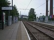 A set of two tram tracks between two platforms with shelters. On the right side there are red and blue flags with "IKEA" written and two industrial chimneys.