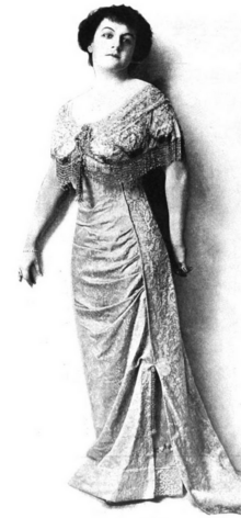 A standing white woman with dark hair, wearing a long gown with a deep scoop neckline and lace trim