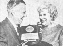 A white man and woman, both smiling, as he presents a plaque to her; the man is wearing a suit and holding the plaque with a hook-like prosthetic hand; the woman has coiffed light hair and is wearing drop earrings and a boatneck sweater