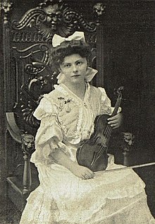 A young white woman, dressed in white with a white bow in her hair, seated in a large, elaborately carved wooden chair, holding a violin.