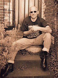 Man sitting on back door steps, wearing jeans, boots, black T-shirt and sun glasses. Rests left elbow on left knee with left forefinger pointing to his right. Right fist hidden inside left hand and he is facing forward.