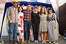 Six people stand in a row with the tallest roughly at the left and the shortest at the right. From left to right: a tall white man with a short black beard, blue jeans, a plaid shirt, and a broad straw hat whose cheeks are made up in red; a white woman with long dark hair in a dress that appears to be made of a quilt; a taller white man with short black hair, a black moustache, and rosy cheeks in a red plaid shirt, blue jeans, and work boots; Fink, a white woman with brown hair and earrings dressed in a black shirt and black jeans; a white woman with two bows in her long brown hair wearing a greenish knee-length floral sundress; and a slightly darker-skinned woman with black hair in a blue checkered dress beneath a blue and white floral vest. Each person pictured has their arms behind the backs or on the shoulders of the person next to them and all are smiling.