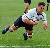 A man wearing a rugby uniform consisting of a white shirt, dark blue shorts, and dark blue socks dives in an attempt to score a try.