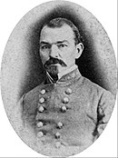 Black and white photo shows a man with a large moustache and a small beard. He wears a gray, double breasted uniform with a general's star on the collar.