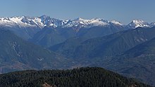 A range of snow-capped mountains. In the foreground is a stretch of forest. In between is an area of lower ground.