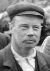 Fuzzy head-and-shoulders photo of a 40-year-old man in a cloth cap and mustache