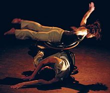 a dancer streched out balances on another dancers wheelchair who is curled over, they are on-stage with theatre lights highlighting the dance