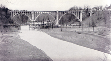 "A black and white photograph of two bridges crossing a valley. The bridge in the background towers over the valley, concrete arches rising high from the ground. The bridge in the foreground is minuscule in comparison, a tiny iron bridge over the creek running through the valley."