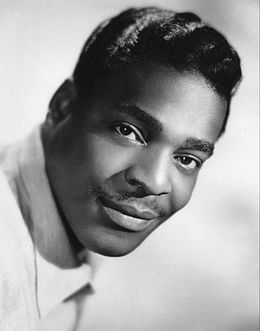 A dark-skinned man with a thin mustache, smiling slightly