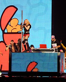 A middle aged blond woman dancing on a DJ's desk wearing red shorts and a black sleeveless shirt. She's holding on to a pole with her left hand and holds a microphone to her mouth with her right hand. She is flanked by dancers