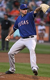 Pitcher in blue baseball cap coming out of his windup as he delivers a pitch to the plate