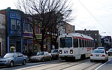 A white single-car trolley in street running. On-street parking combined with double track on a two-lane street leaves limited room for automobile maneuverability.