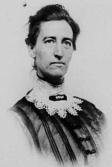 A white woman with dark hair and eyes, and a long neck wrapped in a lace scarf with a brooch at the throat. She is wearing a striped dress. Her hair is loosely drawn back away from her face.