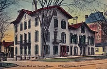 A hand-tinted postcard showing a Victorian-looking white building with many doors and windows and a pointed red roof