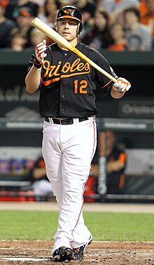 A man, wearing a black batting helmet, a black baseball uniform with the Baltimore Orioles script logo and the uniform number 12 across his chest, clutches his bat with his left hand.