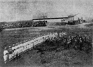 A black and white photograph of a 30 baseball players in light uniforms lined up beside a marching band in dark outfits in the outfield of a baseball park with the grandstand in the background