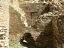 Inside daytime view of a ruined and ceiling-less rectangular room. Tawny-beige stacked sandstone bricks compose walls rising from brush-covered ground. The several walls visible in the image are up to perhaps a dozen feet in height. In the wall immediately at center, a triangle-shaped entrance several feet high leads to an adjacent chamber behind. The upper part of the same wall, shaped like an inverted-triangle, has fallen away or otherwise been removed, revealing a rectangular doorway leading to yet another concealed room. At left and right are two similar walls perpendicular to the one at middle.