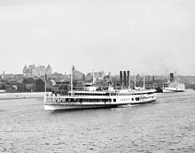 A white steam ship is seen near the shore of the Hudson River in front of the downtown area of Albany; the New York State Capitol can be seen in the background.