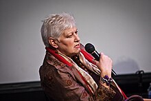 Anne Beatts is facing right, holding a microphone. She has gray hair. She is wearing a scarf and a brown jacket. She has a serious expression.