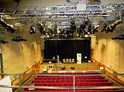 View of a theatre stage from the centre of the balcony. The stage is in the middle of being prepared for an event; there are chairs, microphones and foldback speakers in place. The top of the photograph features the theatre's lighting rig, above the auditorium. The theatre's red stalls seats are visible.