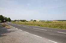 A photo of the airfield, in the foreground is a roadway, with a lay-by on the left hand side. To the right of the image is grass and vegetation, and in the centre of the image off in the distance are a number of buildings.