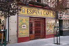 A closed bar façade covered in painted ceramic tiles. On top, it reads "14" and "ESPAÑA CAÑÍ". To the left of the fenced gate, a bullfighter hat in a decorated panel. To the right of the gate, a nude woman, two lovers in Andalusian clothes and other figures.