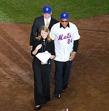Mays in a New York Mets uniform stands on a baseball field and smiles, accompanied by a man in a suit with a New York Mets baseball cap on and a woman in a black dress with a microphone who is speaking
