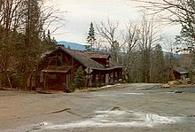 Two unmaintained highways intersect near a large, dilapidated cabin. A second cabin is in the background, reachable by one of the roads. The area is completely surrounded by trees, and large mountains are visible off in the distance.