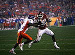 A football player wearing white pants and a red jersey runs with the ball as he tries to deflact a would-be tackler wearing orange pants and white and orange jersey.