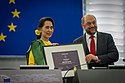 A photograph of two people, a woman on the left and a man on the right, the woman wearing yellow and green and the man wearing a grey lounge suit and a tie