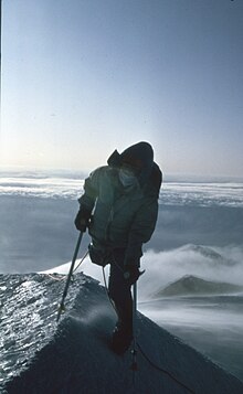 A person with one leg and crutches on a very narrow ridge of snow with clouds and mountain peaks below her