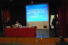 A speaker and two assistants are on stage with a digital presentation displayed on a screen behind them. The slide on the presentation reads: "Great Ideas for Kids from 10 to 12 Years Old: Songs Inspire Delight in Words and Enrapture in Culture".