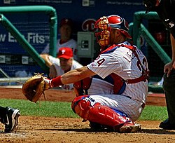 A man in a white baseball uniform with red stripes and wearing a red catcher's mark, red shinguards and cleats, and a light-brown catcher's mitt